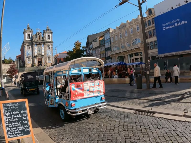 Ulična scena u Portu, Portugal, sa svetloplavim tuk-tuk vozilom u prvom planu koje vozi turiste. U pozadini se vidi crkva svetog Ilije sa prepoznatljivim fasadama ukrašenim azulejos pločicama. S desne strane su pešaci, lokalne prodavnice i tabla sa natpisom "Reabilitação do Cinema Batalha". Atmosfera je sunčana i živa.