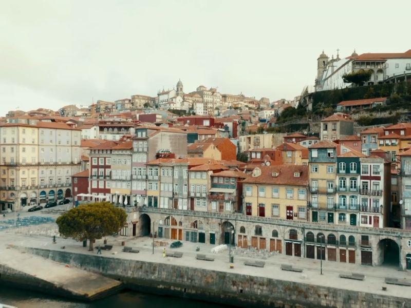 Pogled na šareni stari deo Porta sa tradicionalnim kućama koje imaju crvene krovove i fasade u raznim bojama. Ispred kuća nalazi se šetalište uz reku Douro, dok se grad prostire uz blagi nagib uzbrdo sa crkvama i drugim istorijskim zgradama na vrhu. Nebo je oblačno, a atmosfera je mirna.