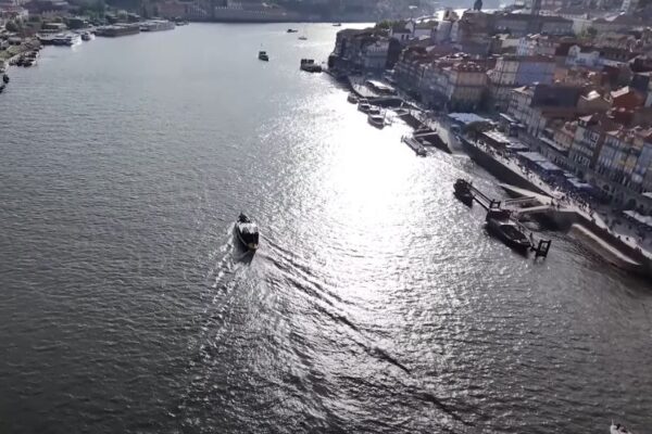 Pogled iz vazduha na reku Douro u Portu, Portugal. Na desnoj strani reke nalazi se istorijski deo grada Ribeira, sa šarenim zgradama uz obalu. Nekoliko čamaca i brodova plovi rekom, a sunčeva svetlost se reflektuje na površini vode.