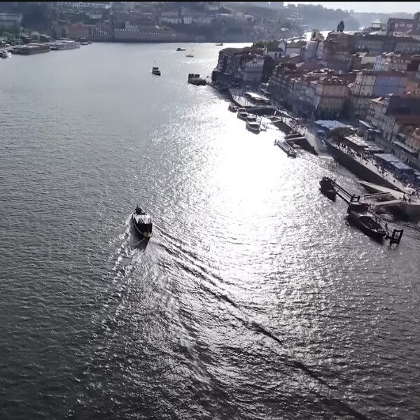 Pogled iz vazduha na reku Douro u Portu, Portugal. Na desnoj strani reke nalazi se istorijski deo grada Ribeira, sa šarenim zgradama uz obalu. Nekoliko čamaca i brodova plovi rekom, a sunčeva svetlost se reflektuje na površini vode.