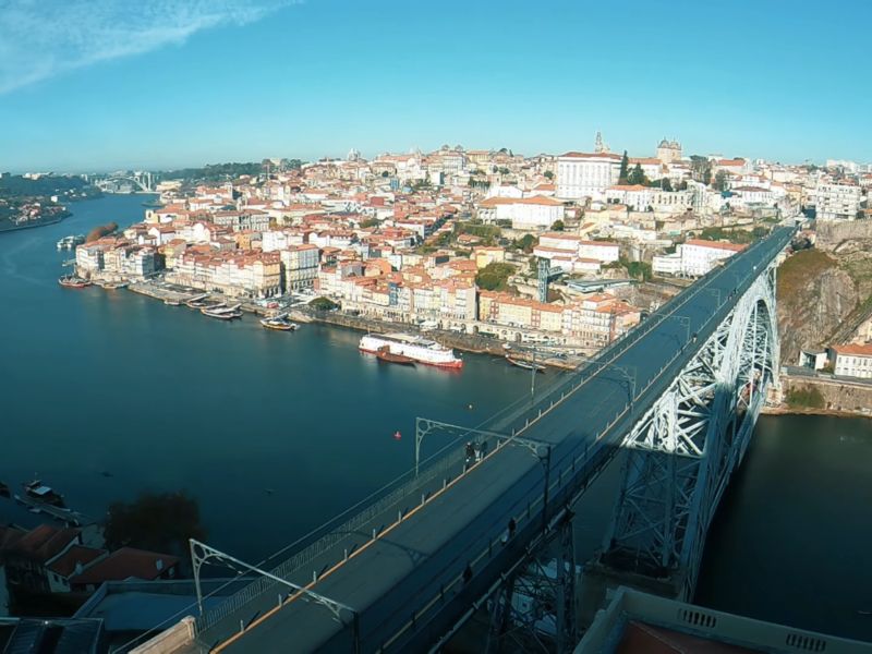 Pogled iz vazduha na most Dom Luís I koji prelazi reku Douro u Portu, Portugal. S desne strane mosta pruža se istorijski deo grada Ribeira, prepoznatljiv po šarenim kućama i uskim ulicama. Na reci su usidreni čamci, a nebo je vedro i plavo.