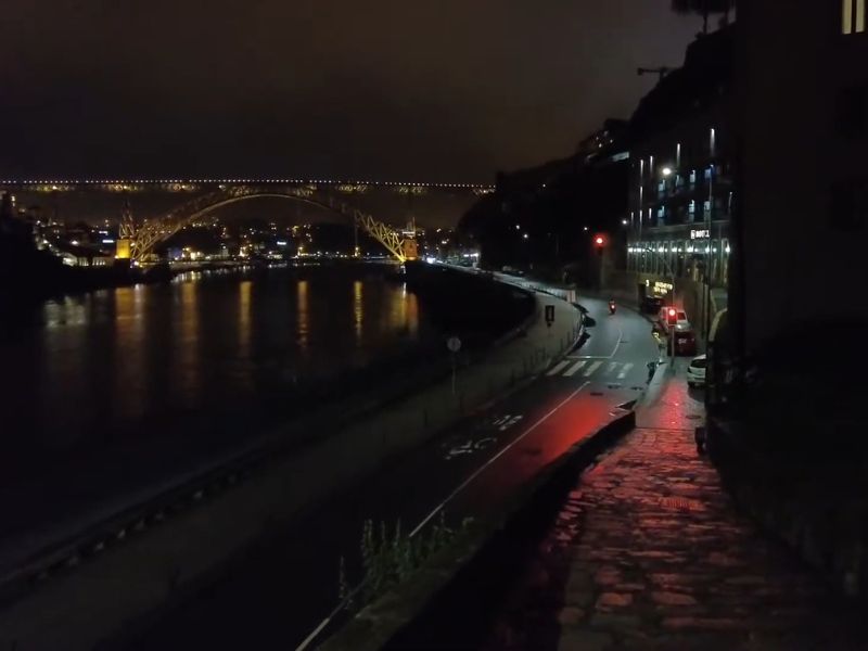 Noćni prizor iz Porta, Portugal, sa osvetljenim mostom Dom Luís I u daljini koji prelazi preko reke Douro. Ulica uz obalu je slabo prometna, a svetla restorana i uličnih lampi reflektuju se na mokrom kolovozu. Atmosfera je mirna i romantična.