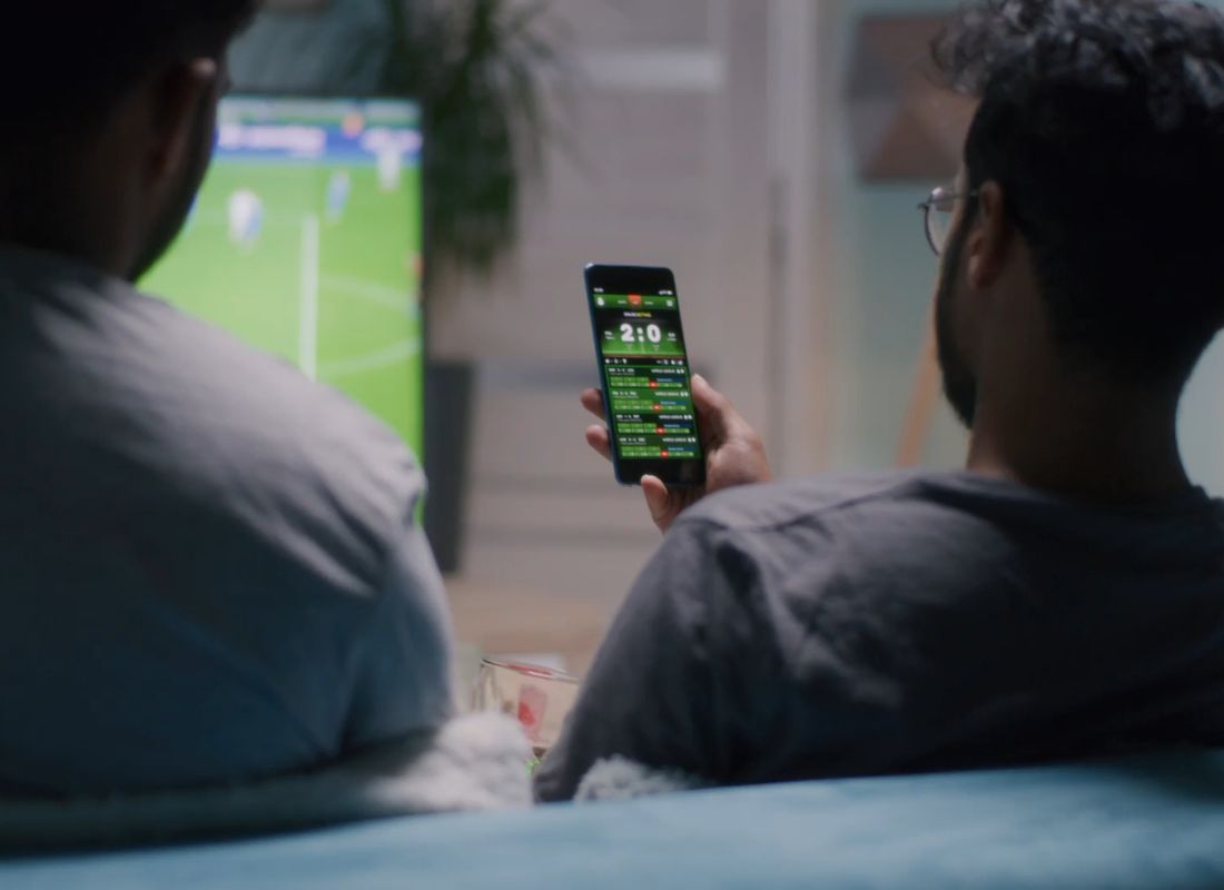 dva momka sede na ugaonoj, gledaju fudbal na televizoru i proveravaju rezultate utakmice na telefonu