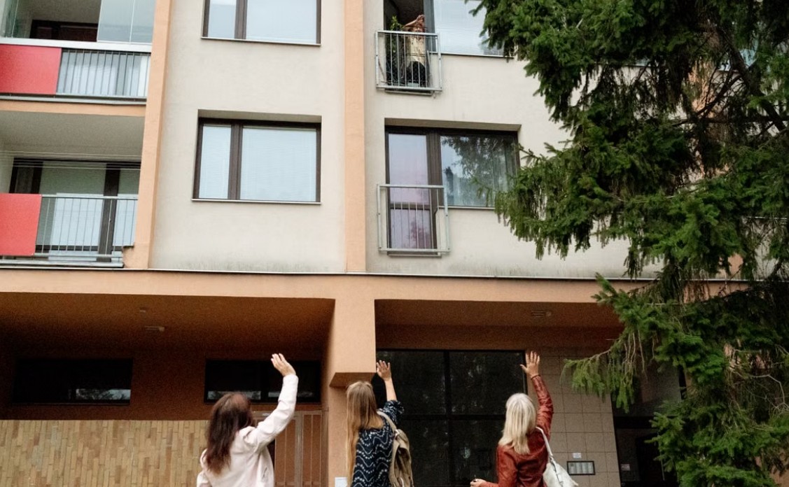 Tri devojke sa podignutim rukama stoje ispred višespratne stambene zgrade sa balkonima i velikim prozorima