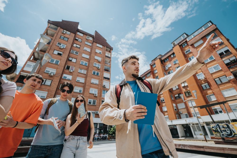 Grupa studenata sa rancima i sveskama stoji ispred višespratnih stambenih zgrada, dok mladić u prvom planu pokazuje rukom u daljinu