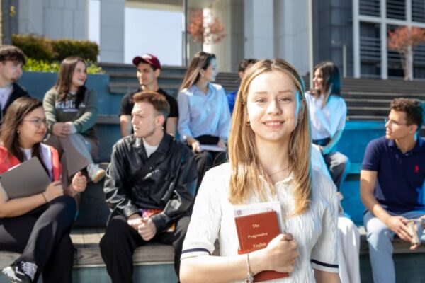 Grupa studenata sedi na stepenicama ispred fakultetske zgrade, dok devojka u prvom planu drži knjigu i gleda u kameru