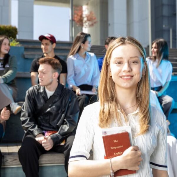 Grupa studenata sedi na stepenicama ispred fakultetske zgrade, dok devojka u prvom planu drži knjigu i gleda u kameru