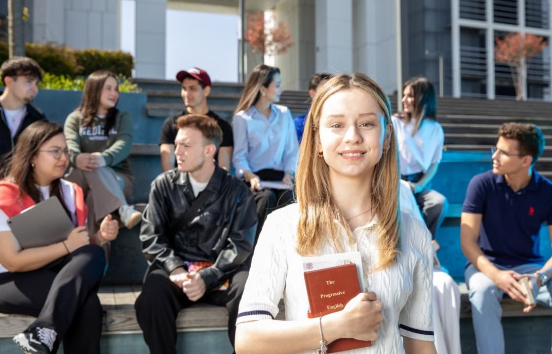 Grupa studenata sedi na stepenicama ispred fakultetske zgrade, dok devojka u prvom planu drži knjigu i gleda u kameru