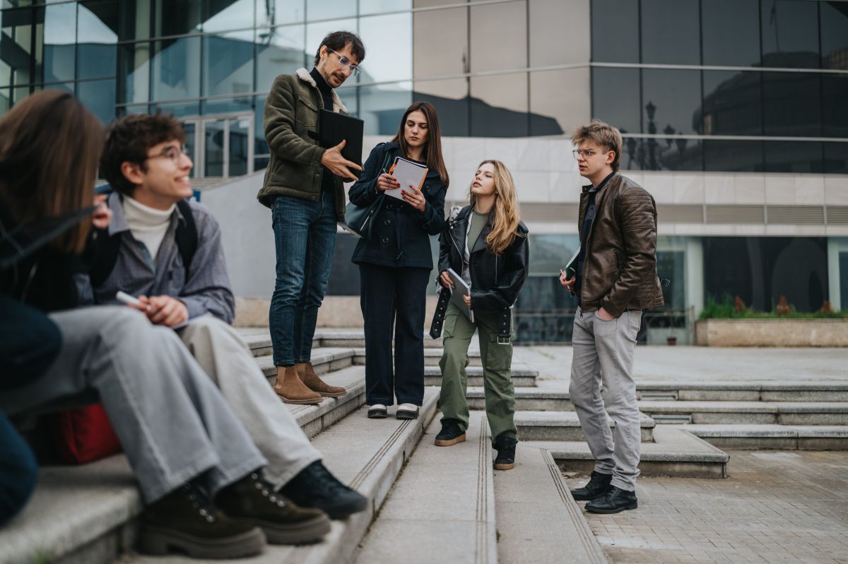 Grupa studenata sa sveskama i laptopom razgovara na stepenicama ispred moderne fakultetske zgrade